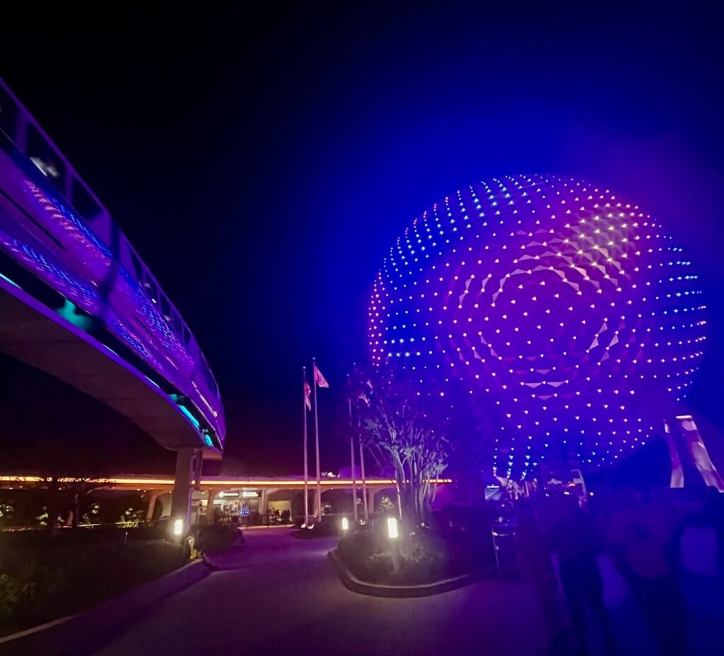 A nighttime shot of the monorail passing by next to a lit up Spaceship Earth with purple lights with minimal crowds at EPCOT's entrance.