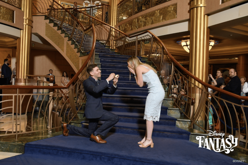 Picture taken with Disney Cruise Line's Unlimited Photo Package of a young man proposing to his girlfriend in front of the grand staircase aboard a Disney Cruise ship.