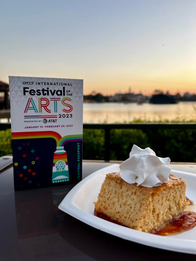 Image of a 2023 Festival of the Arts booklet and coconut flan dessert in front of World Lagoon during sundown.