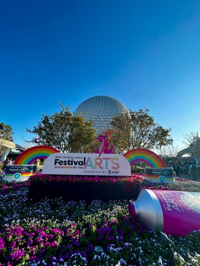 Image of a sunny day at EPCOT of the Festival of the Arts display with Spaceship Earth in the background. The display consists of a massive festival sign, rainbows and a paint tube on the manicured garden.