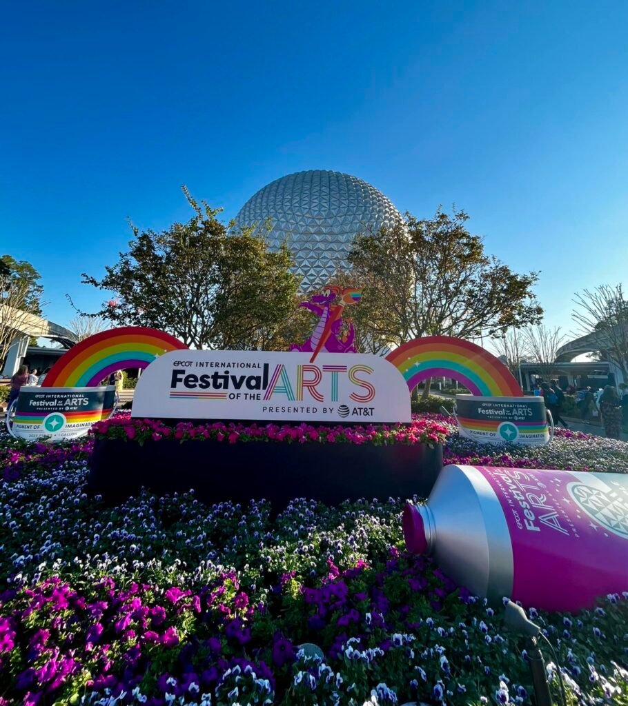 Image of a sunny day at EPCOT of the Festival of the Arts display with Spaceship Earth in the background. The display consists of a massive festival sign, rainbows and a paint tube on the manicured garden.