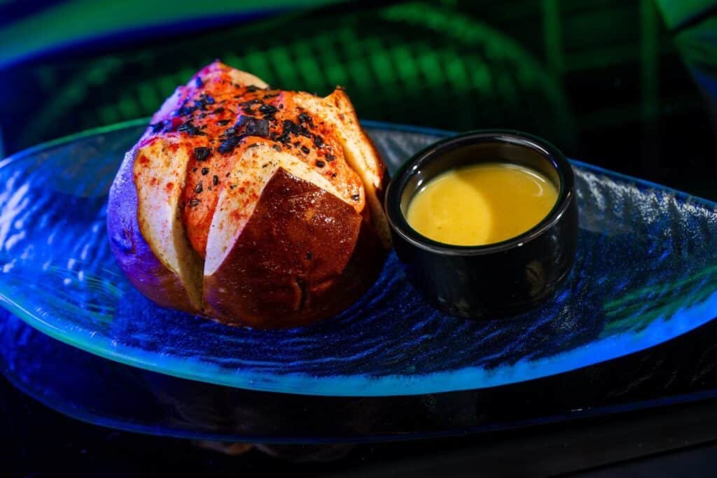 Disney's image of the. Umbaran Cheese Roll which is a spice-dusted pretzel bun stuffed with herbed cheese and dipping sauce on the side.