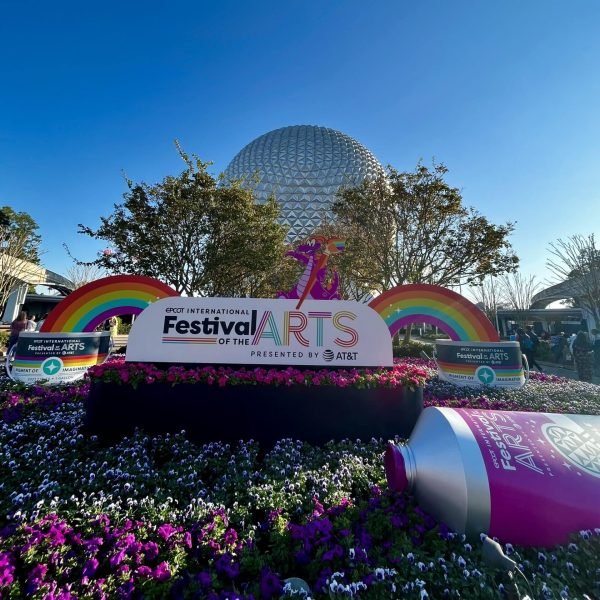 Image of a sunny day at EPCOT of the Festival of the Arts display with Spaceship Earth in the background. The display consists of a massive festival sign, rainbows and a paint tube on the manicured garden.