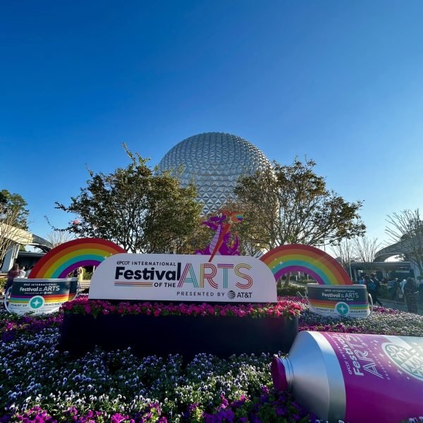 Image of a sunny day at EPCOT of the Festival of the Arts display with Spaceship Earth in the background. The display consists of a massive festival sign, rainbows and a paint tube on the manicured garden.