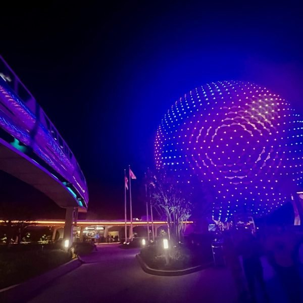 A nighttime shot of the monorail passing by next to a lit up Spaceship Earth with purple lights with minimal crowds at EPCOT's entrance.